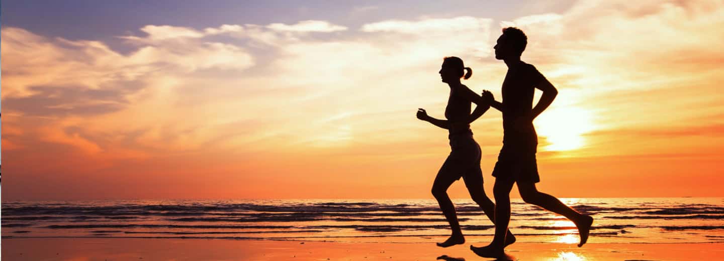 Silhouettes of a man and woman jogging on the beach at sunset, showing how to stay fit while traveling, waves and sky behind them.