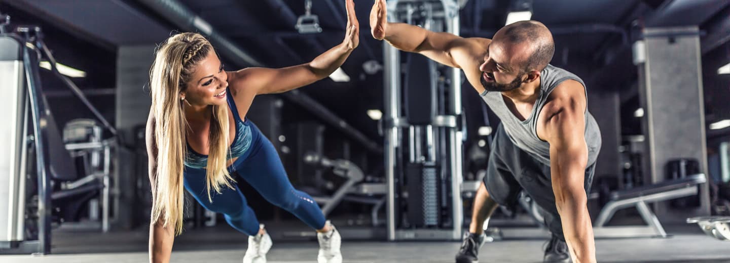 Two travelers in gym attire do side planks and high-five at a hotel fitness center, showing it's easy to stay fit while on the road.