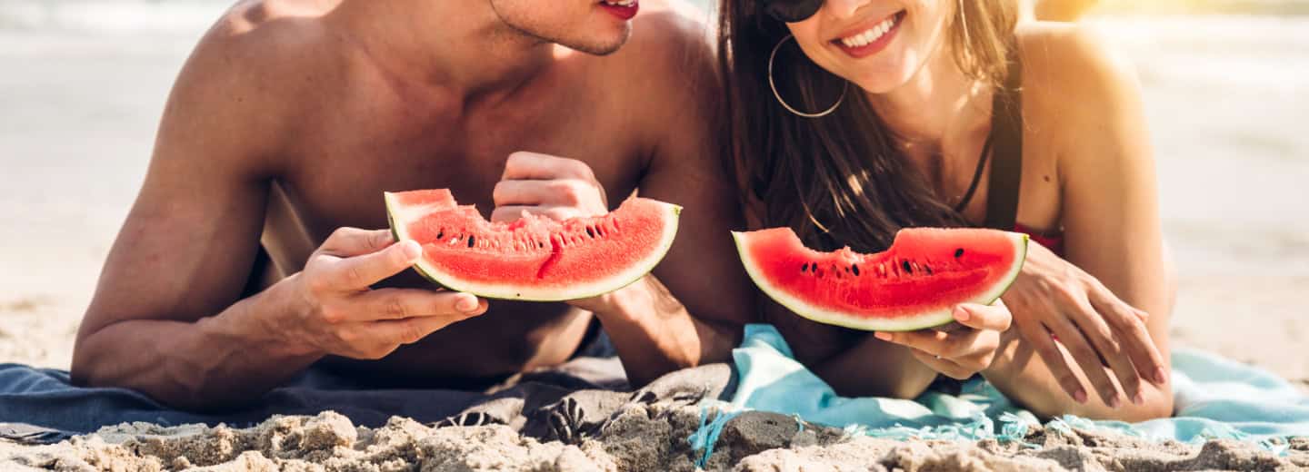 A couple sits on the beach, smiling and enjoying watermelon slices as a healthy snack to stay energized while traveling.