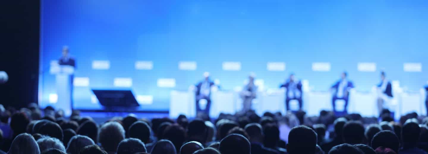 Travelers at a conference watch speakers on stage, highlighting ways to stay fit while on the go.