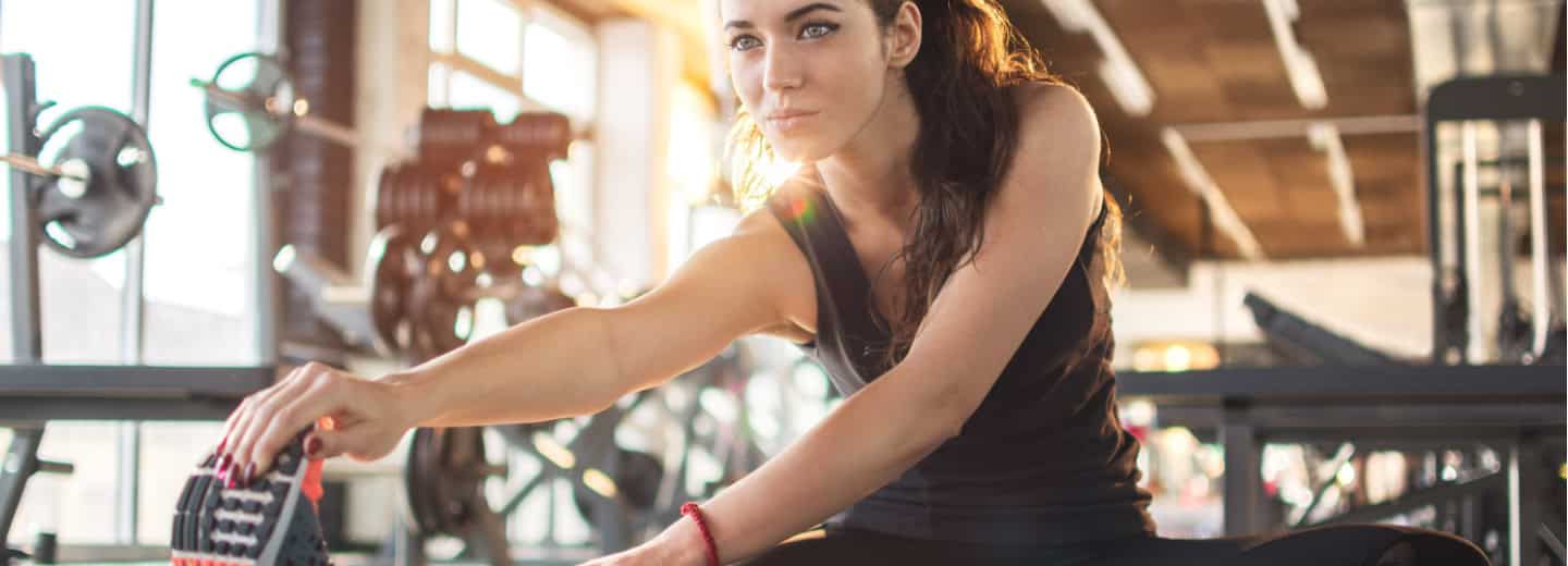 Woman in gym clothes does a warm-up stretch on the floor of a bright gym, showing how to stay fit while traveling.