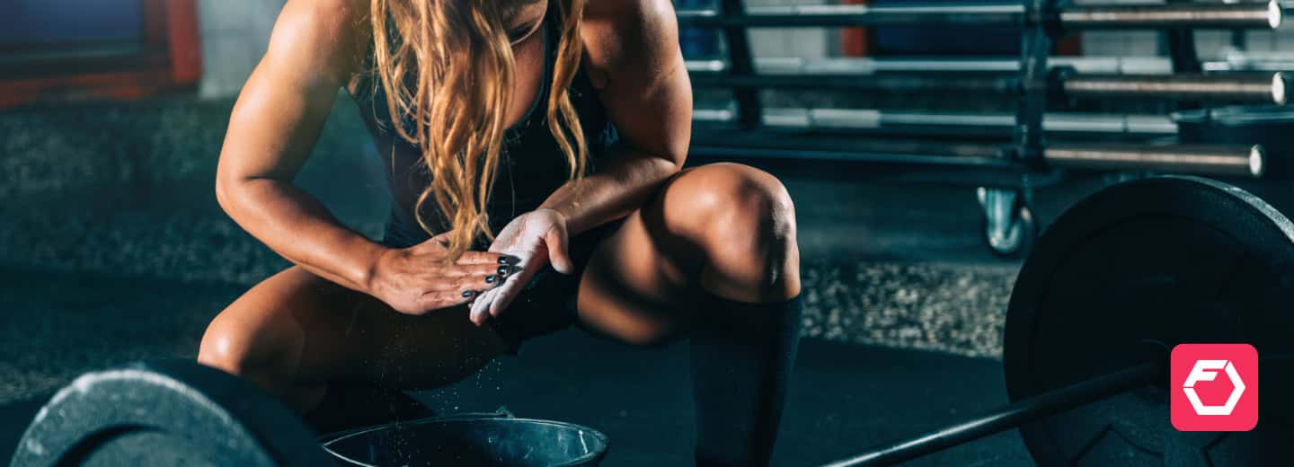 A woman prepares to lift a barbell by chalking her hands in a well-equipped gym, illustrating how travelers can maintain their fitness routines on the go. The HotelGyms.com logo is visible in the lower right corner.