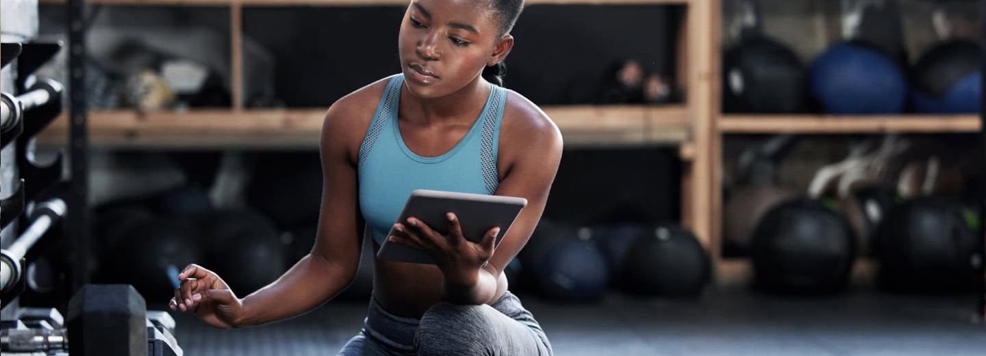 Woman in athletic wear checks her workout plan on a tablet, kneeling by gym equipment in a hotel fitness studio for travelers. Helping to get a GymFactor score.