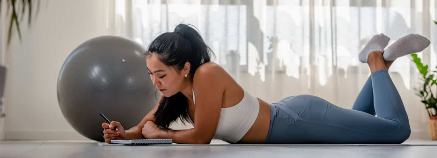Woman writes her new years resolution in a notebook on the floor of her hotel room, exercise ball and plants nearby for an active stay.