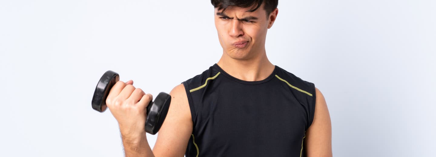 Man lifts a dumbbell in a hotel gym, appearing unsure how to use the equipment while traveling.