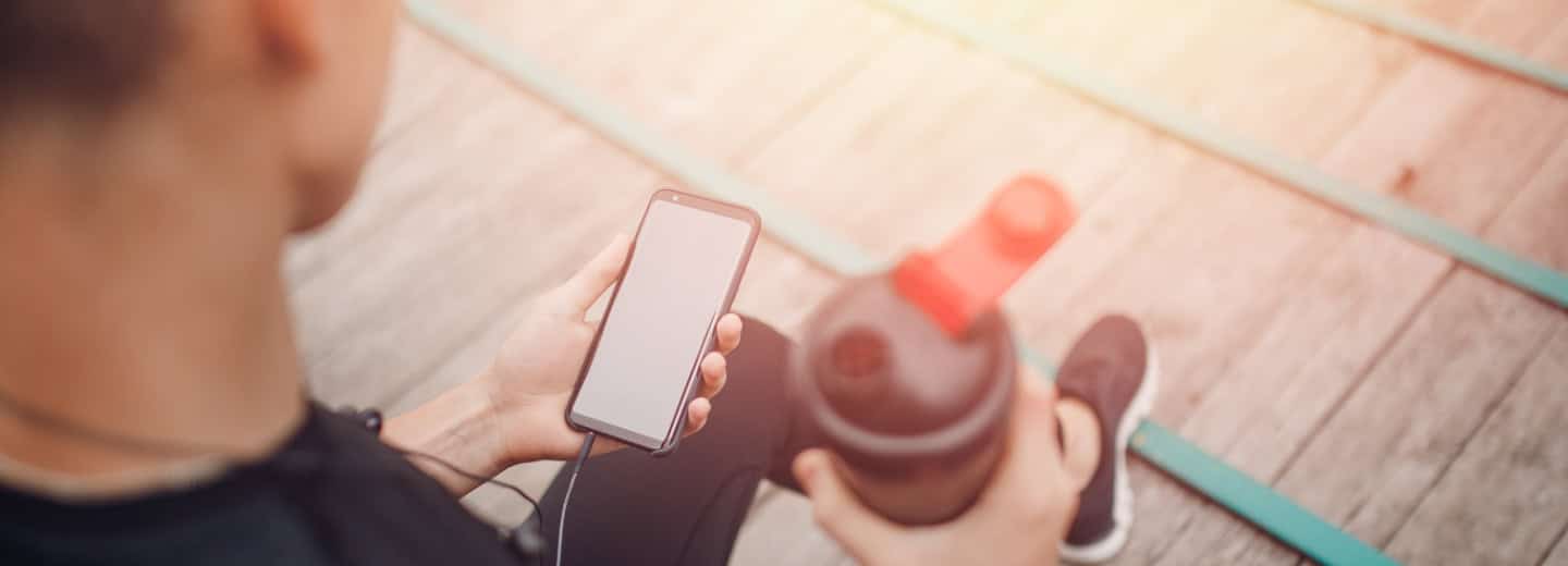 Traveler in athletic wear sits outside, holding a smartphone and shaker bottle, staying active while away from home.