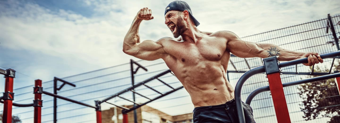 Man flexing biceps and smiling at an outdoor workout park, showing how to stay fit while traveling, wearing a backward cap.