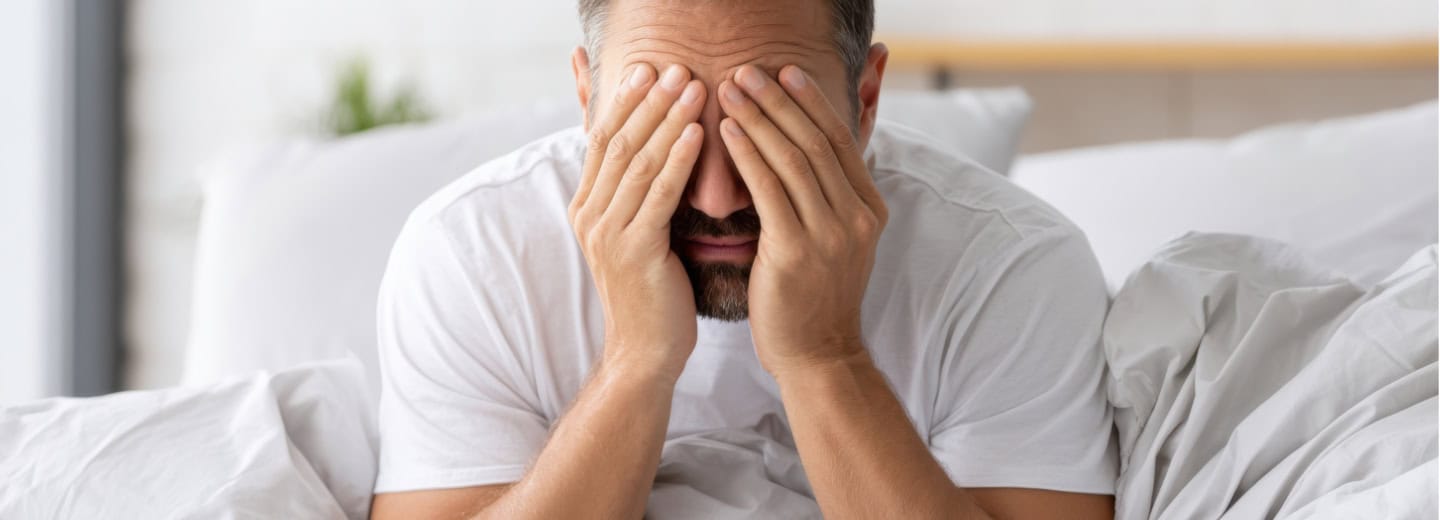 A man sits on a hotel bed, covering his face with his hands, looking tired or stressed, possibly from missing a workout while traveling.