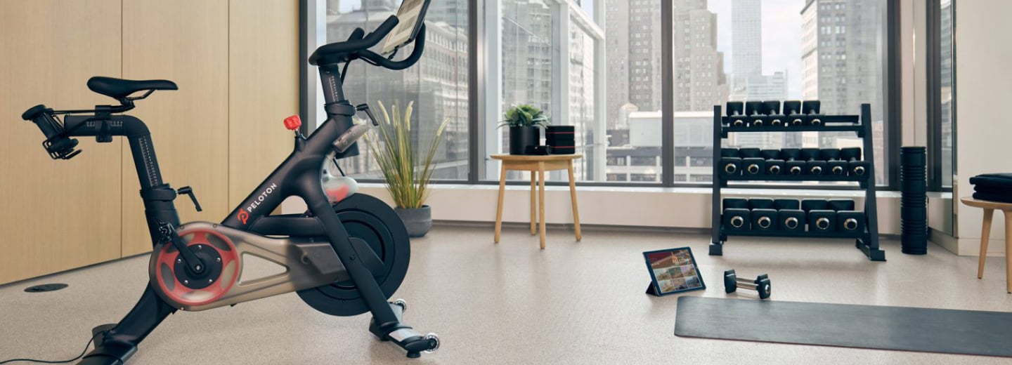 Stay fit while traveling in a hotel gym featuring a Peloton bike, dumbbells, yoga mat, and large windows with city views.