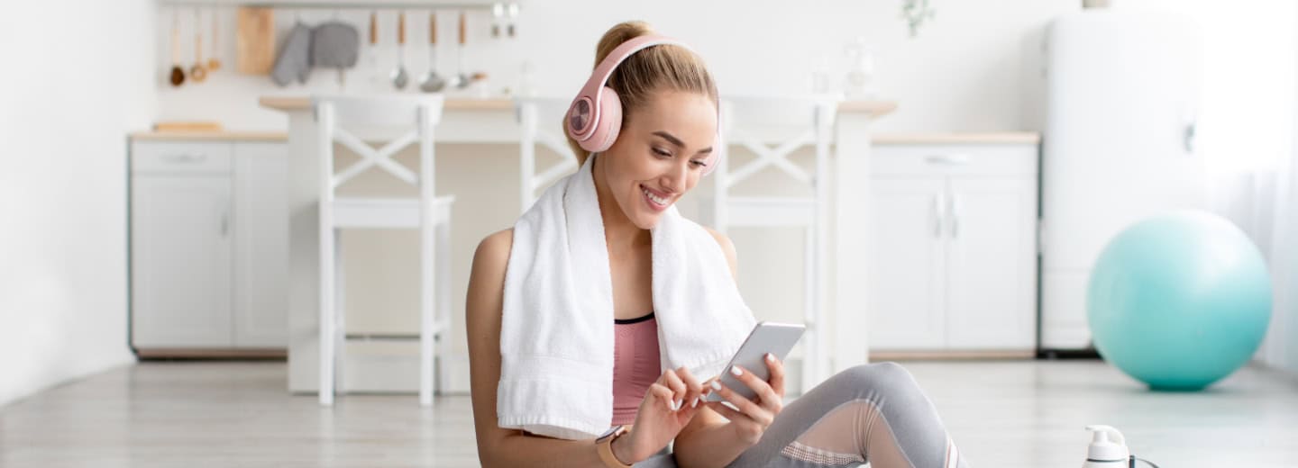 Woman in workout clothes and headphones sits on the floor of a bright kitchen, using Apple Fitness+.