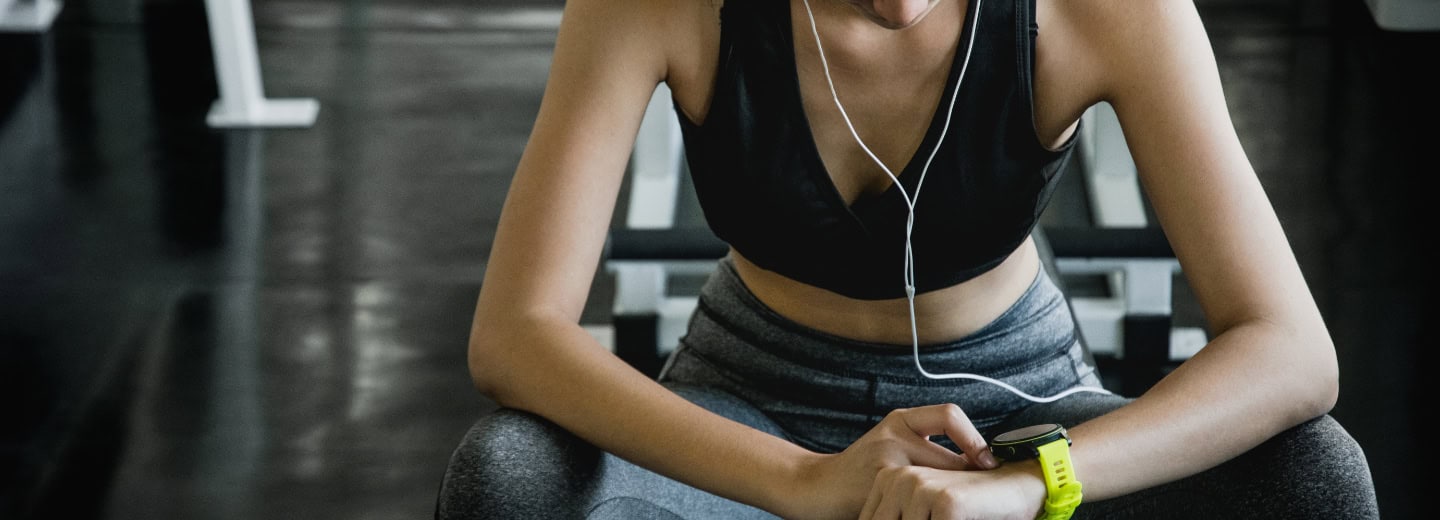 Woman in athletic wear sits on a gym bench, wearing earphones and checking her smartwatch, showing how to stay fit while traveling.