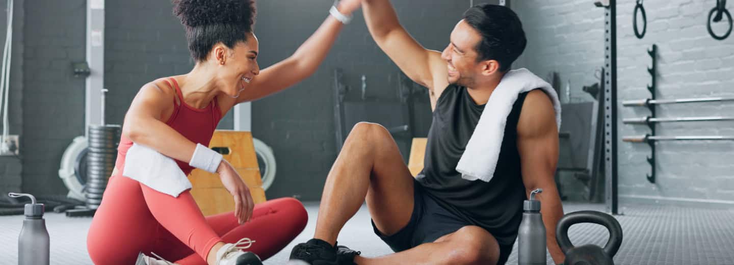 Two travelers in workout clothes sit on a hotel gym floor, smiling and high-fiving after staying active during their trip.