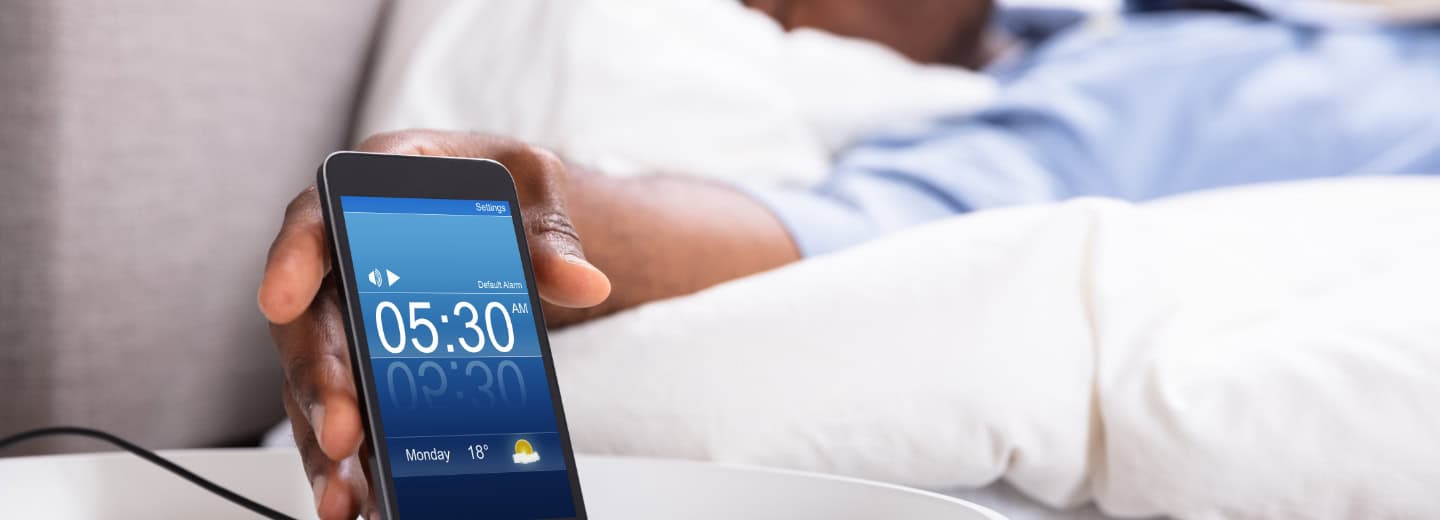 Traveler in bed grabs a phone showing a 5:30 AM alarm, emphasizing an early start to stay active while on the road.