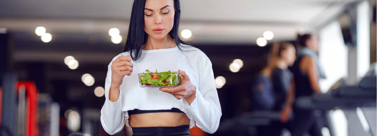 Traveler in gym attire enjoys a salad at a hotel gym, with others working out nearby, highlighting healthy habits on the go.