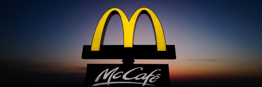 Bright yellow McDonald's arches and a McCafé sign glow against a dark sunset sky, marking a quick meal stop for travelers staying fit.