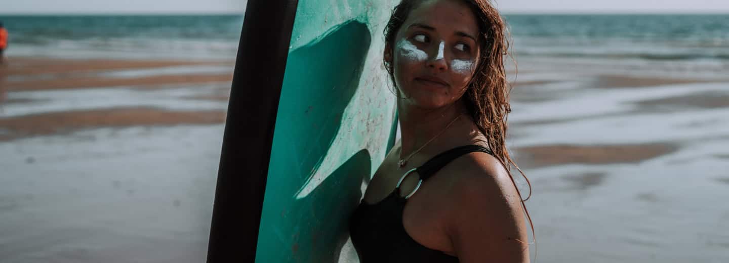 Woman with sunscreen on her face holds a surfboard at the beach, staying active and fit while traveling.
