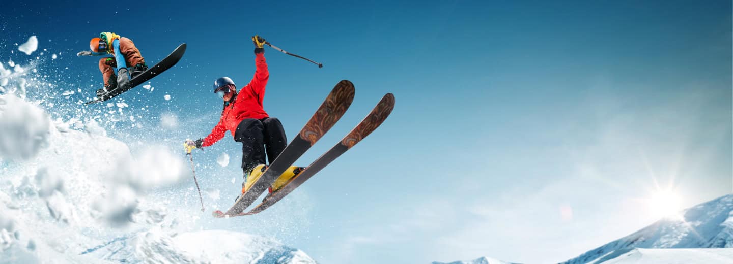 A skier and a snowboarder catch air on snowy slopes under a clear blue sky, showing an active way to stay fit while traveling.