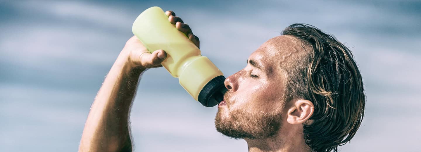 Traveler staying hydrated after a workout outside, drinking from a yellow bottle under a blue sky, sweat visible on his face.