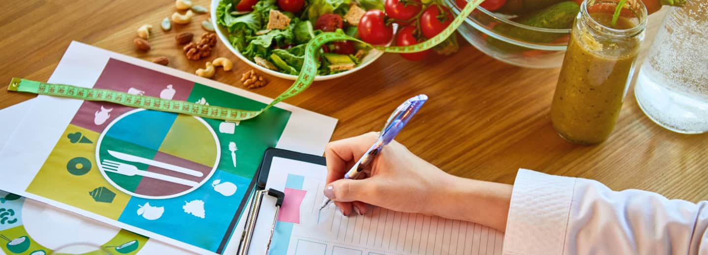 A person creates meal plans at a desk with healthy food, charts, and a measuring tape to stay fit while traveling.