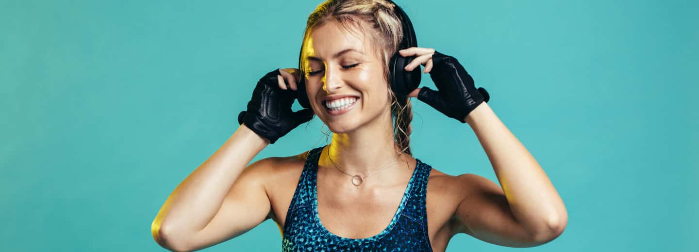 A woman in workout clothes and gloves enjoys music on headphones, ready to stay fit while traveling, teal background behind her.