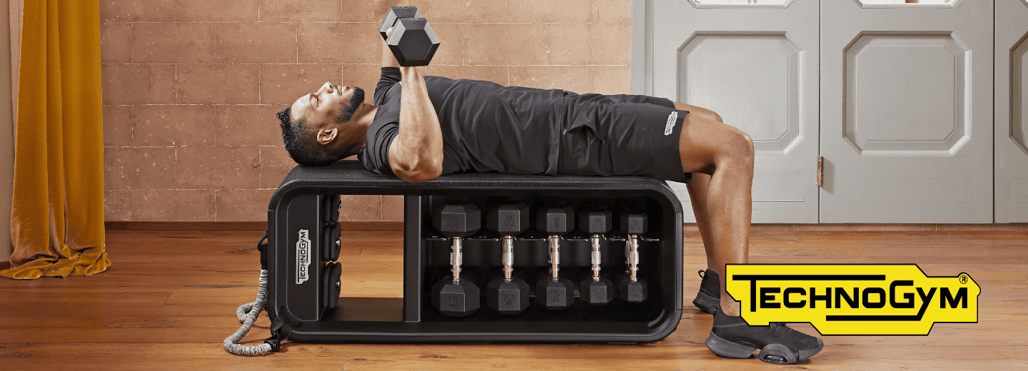 Traveler working out on a Technogym bench in a hotel gym, lifting a dumbbell with additional dumbbells stored underneath.