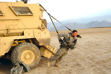 A soldier in uniform stays fit while traveling by pulling a military vehicle with a harness across a desert, mountains behind.